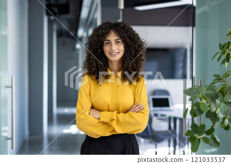 Confident businesswoman with curly hair in yellow blouse stands with arms crossed in modern office hallway. Strong, accomplished professional representing leadership, success, empowerment. Confident businesswoman with curly hair in yellow blouse stands with arms crossed in modern office hallway. Strong, accomplished professional representing leadership, success, empowerment. 121033537