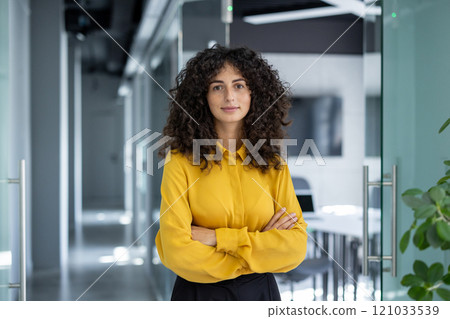 Confident businesswoman standing with arms crossed in modern office hallway. Image conveys professionalism, leadership, and success in corporate environment. Bright and inspiring scene. 121033539