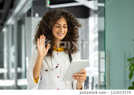 A smiling medical professional engages in a virtual consultation using tablet. The doctor, with a stethoscope around her neck, waves she interacts online, showcasing approach to healthcare. 121033574