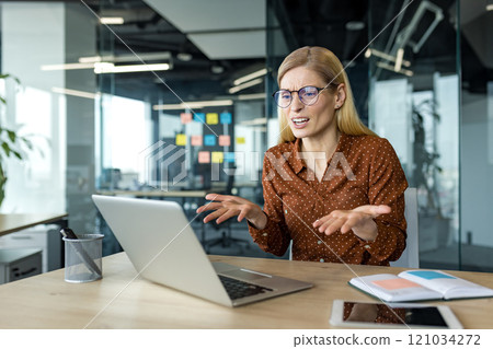 Mature business woman displays frustration during online conversation in office setting. Laptop and open notebook accentuate professional vibe, emphasizing challenges of remote communication. 121034272