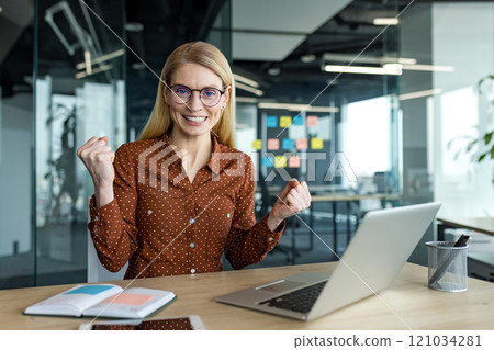 Mature business woman celebrating success in modern office, seated at desk with laptop open, raising fists in excitement. Confident expression represents achievement and positive progress. Mature business woman celebrating success in modern office, seated at desk with laptop open, raising fists in excitement. Confident expression represents achievement and positive progress. 121034281