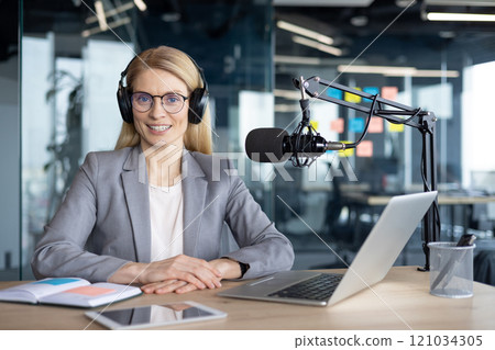 Mature businesswoman using microphone for podcast recording in modern office. Headphones and laptop on desk suggest professional setup for broadcasting or virtual communication. 121034305