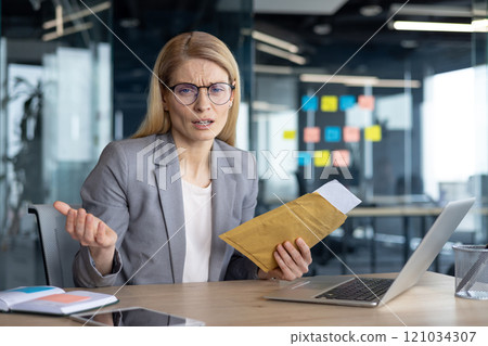 A perplexed businesswoman in a gray suit holding an envelope at a desk, engaging with a laptop in a contemporary office setting, possibly amidst a discussion or meeting. A perplexed businesswoman in a gray suit holding an envelope at a desk, engaging with a laptop in a contemporary office setting, possibly amidst a discussion or meeting. 121034307