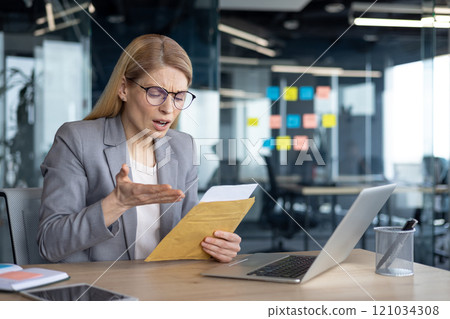 A mature businesswoman sits at her desk reading an envelope with a surprised expression. Her work environment includes a laptop and colorful sticky notes on the glass walls. 121034308