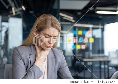 A businesswoman in a suit holds her throat, showing discomfort, in a contemporary office. Her expression indicates pain or stress, capturing a moment of vulnerability in a professional environment. A businesswoman in a suit holds her throat, showing discomfort, in a contemporary office. Her expression indicates pain or stress, capturing a moment of vulnerability in a professional environment. 121034322