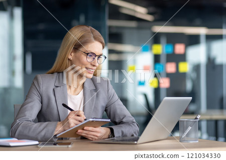 A professional woman in a suit is using a laptop in a modern office, jotting down notes with a smile, showcasing confidence and productivity during a meeting or work session. 121034330
