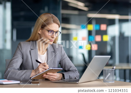 Mature woman businesswoman working diligently on a laptop, taking notes with focus. This professional environment highlights her dedication and expertise. Ideal for business and office themes. 121034331