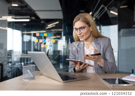 Mature woman businesswoman in office having virtual meeting using laptop and notebook for notes. Professional interaction, online collaboration, productivity in modern workspace. 121034332