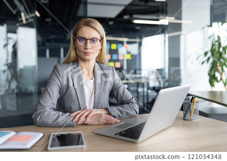 A professional businesswoman sits confidently at her desk in a modern office setting, equipped with a laptop and smartphone, reflecting a blend of technology and professionalism. 121034348
