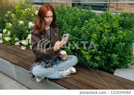 Charming young woman in leather jacket enjoying coffee break sitting on city bench, checking phone. Pretty redhead female using smartphone holding coffee outdoors. Charming young woman in leather jacket enjoying coffee break sitting on city bench, checking phone. Pretty redhead female using smartphone holding coffee outdoors. 121034560