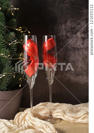 Christmas decor, two transparent glass with a red ribbon inside, tree with lights, dark background, beige cloth on wooden table 121035112