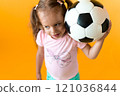 Authentic cute smiling preschool little girl with classic black and white soccer ball look at camera on yellow background. child play football in t-shirt and shorts. Sport, championship, team concept 121036844