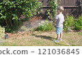 Funny little boy watering lawn plants in garden housing backyard. Adorable child playing with irrigation hose at hot sunny summer outdoors. Children help with housework. activity for kids. Childhood 121036845