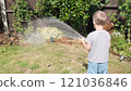 Funny little boy watering lawn plants in garden housing backyard. Adorable child playing with irrigation hose at hot sunny summer outdoors. Children help with housework. activity for kids. Childhood 121036846