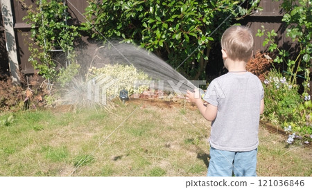 Funny little boy watering lawn plants in garden housing backyard. Adorable child playing with irrigation hose at hot sunny summer outdoors. Children help with housework. activity for kids. Childhood 121036846