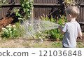 Funny little boy watering lawn plants in garden housing backyard. Adorable child playing with irrigation hose at hot sunny summer outdoors. Children help with housework. activity for kids. Childhood 121036848