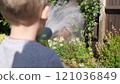 Funny little boy watering lawn plants in garden housing backyard. Adorable child playing with irrigation hose at hot sunny summer outdoors. Children help with housework. activity for kids. Childhood 121036849