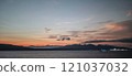 A tranquil scene of the sea at dawn with soft hues of orange and blue blending over distant mountains and a calm horizon. 121037032