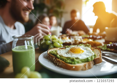 Friends engage in lively conversation over a delightful brunch spread, featuring creamy avocado toast with a sunny-side-up egg, fresh fruits, and nutritious smoothies. 121037293
