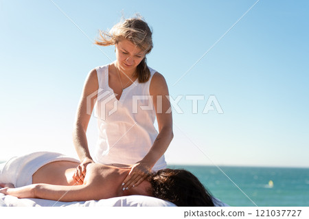 Therapist massaging woman's naked back against sea Therapist massaging woman's naked back against sea 121037727