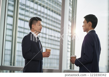 two asian business men standing and talking by the window in office two asian business men standing and talking by the window in office 121038342
