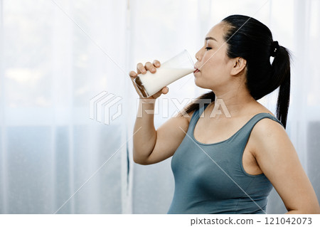 Pregnant woman in gray sleeveless top drinking glass of milk, standing in softly lit room with calm and healthy lifestyle concept 121042073