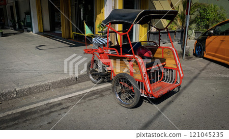 pedicab or rickshaw or locals calls it becak with a canopy is parked on a sunny street, reflecting a vibrant urban scene. The vehicle features a padded seat and unique design pedicab or rickshaw or locals calls it becak with a canopy is parked on a sunny street, reflecting a vibrant urban scene. The vehicle features a padded seat and unique design 121045235