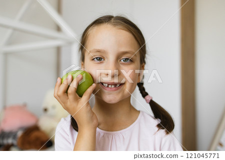 Little Caucasian girl with missing teeth from falling out milk teeth smiling and holding apple indoors, conveying joy and childhood innocence. Little Caucasian girl with missing teeth from falling out milk teeth smiling and holding apple indoors, conveying joy and childhood innocence. 121045771