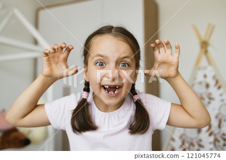 Portrait of playful little Caucasian girl with braided hair and gaps from missing milk teeth posing at home with frightening expression. 121045774