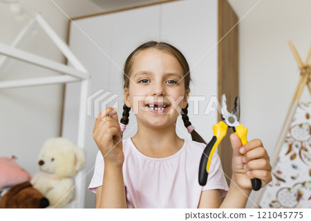 Little Caucasian girl joyfully displays fallen baby tooth with pliers in bright home environment. Celebrating milestone with a cheerful expression. 121045775