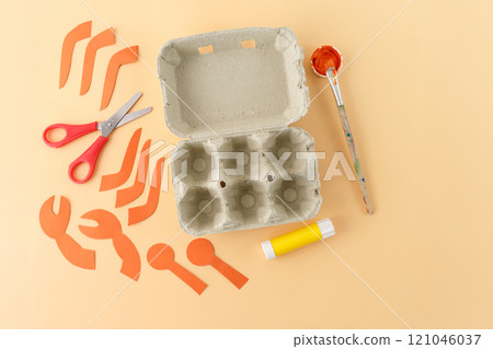 a collection of crafting materials arranged on a light orange surface. egg carton, scissors, glue stick, pieces of orange paper cut, simple and creative DIY project using everyday materials 121046037