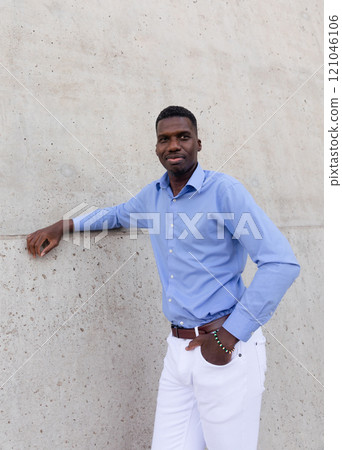 Portrait Of Handsome Smiling Tall African American Man wearing White Jeans and Blue Shirt, Brown Belt Outdoor, Concrete Wall on Background. Copy Space Fashionable Male, Ivorian. Vertical Plane 121046106