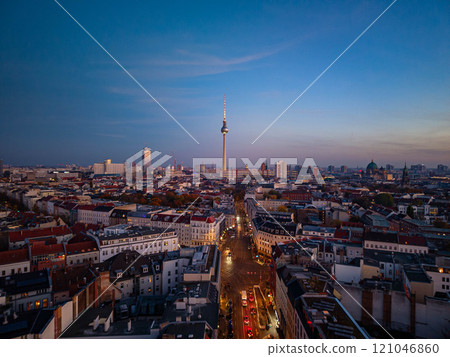 Berlin TV tower, a striking landmark, looms over the cityscape at dusk, illuminating the urban skyline with a warm glow from an aerial view 121046860