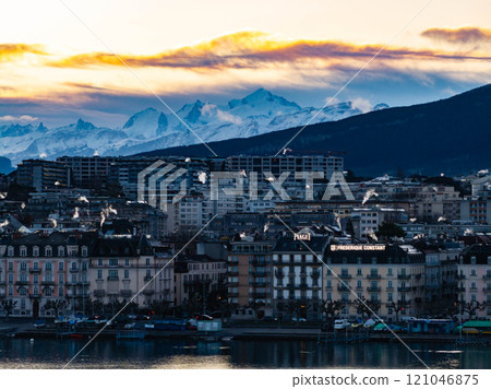 Geneva's cityscape at sunrise features smoking chimneys, waterfront buildings, and Mont Blanc mountain range in the Alps, creating a breathtaking winter scene 121046875