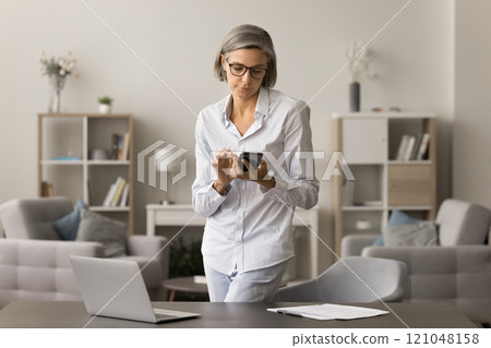 Focused middle aged entrepreneur woman using mobile phone at home Focused middle aged entrepreneur woman using mobile phone at home 121048158