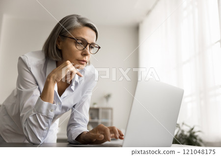 Thoughtful grey haired freelancer woman in glasses looking at laptop Thoughtful grey haired freelancer woman in glasses looking at laptop 121048175