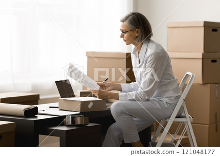 Serious storage employee woman reading parcel card at workplace Serious storage employee woman reading parcel card at workplace 121048177