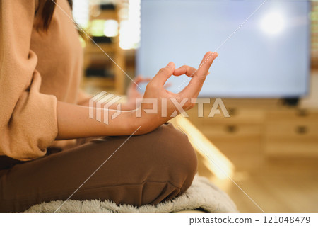 Close up of woman sitting in a lotus position practicing meditation in living room 121048479