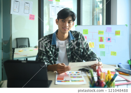 Confident young designer working in modern office surrounded by tools and sketches for UI UX design 121048488