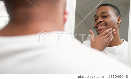 Skin routine. Facial care. Wellbeing freshness. Satisfied smiling man touching face stubble applying moisturizing shea butter looking in mirror on light free space background. Skin routine. Facial care. Wellbeing freshness. Satisfied smiling man touching face stubble applying moisturizing shea butter looking in mirror on light free space background. 121049048
