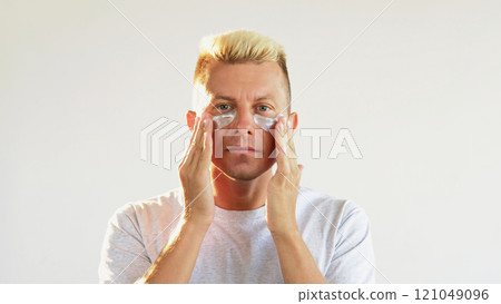 Eye patches. Anti-aging treatment. Confident blond man applying moisturizing collagen hydrogel pads on clean fresh face skin isolated on white empty space. Eye patches. Anti-aging treatment. Confident blond man applying moisturizing collagen hydrogel pads on clean fresh face skin isolated on white empty space. 121049096