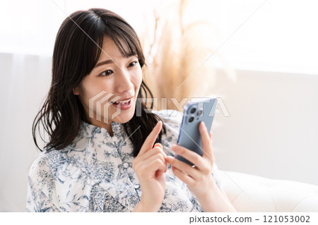 A young woman with long black hair sitting on a white sofa and holding a smartphone 121053002