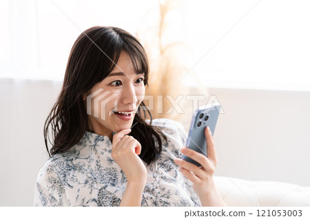 A young woman with long black hair sitting on a white sofa and holding a smartphone 121053003