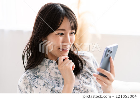 A young woman with long black hair sitting on a white sofa and holding a smartphone A young woman with long black hair sitting on a white sofa and holding a smartphone 121053004