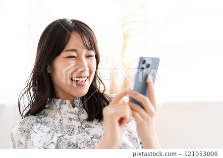 長黑髮的年輕女子坐在白色沙發上,手上拿著智慧型手機 長黑髮的年輕女子坐在白色沙發上,手上拿著智慧型手機 121053008