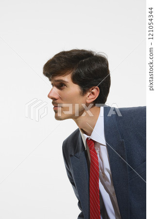 Close-up side profile of businessman in blue suit and red tie, looking ahead with determination against clean white background. 121054344