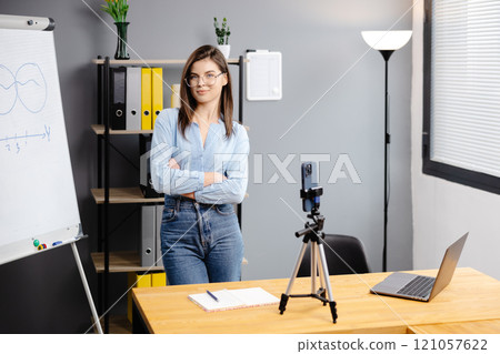Young woman mentor recording presentation, webinar on smartphone standing on tripod, teacher coach explaining, holding online lesson, using phone webcam. Young woman mentor recording presentation, webinar on smartphone standing on tripod, teacher coach explaining, holding online lesson, using phone webcam. 121057622