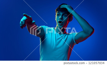 Portrait of handsome young man in checkered shirt emotionally pointing towards with finger against blue studio background in neon light. Announcement, sales 121057834