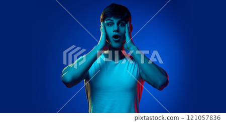 Portrait of handsome young guy in white t-shirt posing with shocked and excited face against blue studio background in neon light 121057836