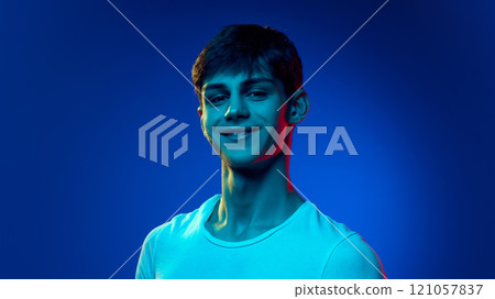 Portrait of handsome young man in white t-shirt posing with smile against blue studio background in neon light 121057837